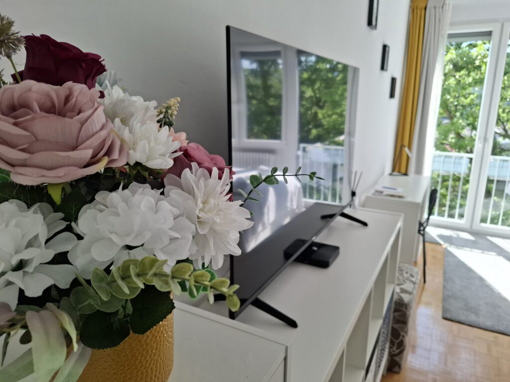 Uređeni i čisti dnevni boravak apartmana s modernim televizorom, svježim cvijećem i pogledom na balkon s prirodnim svjetlom